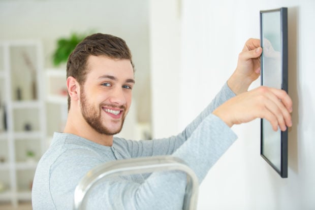 This is a happy man positioning a picture frame on a wall (Photo via Shutterstock)